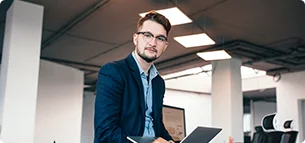 attractive man glassess is sitting near workplace office he wears blue shirt dark jacket he holds laptop looks camera 1